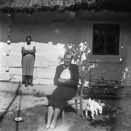 kobieta siedząca na krześle na tle wiejskiej chaty krytej strzechą, pod ścianą domu stojąca druga kobieta, na podwórku przechadza się biały piesek