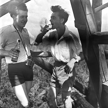 dwaj młodzi mężczyźni piją alkohol, w tle fragment konstrukcji drewnianej i pole uprawne