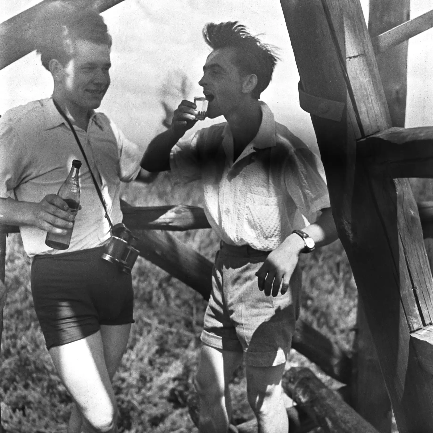 dwaj młodzi mężczyźni piją alkohol, w tle fragment konstrukcji drewnianej i pole uprawne
