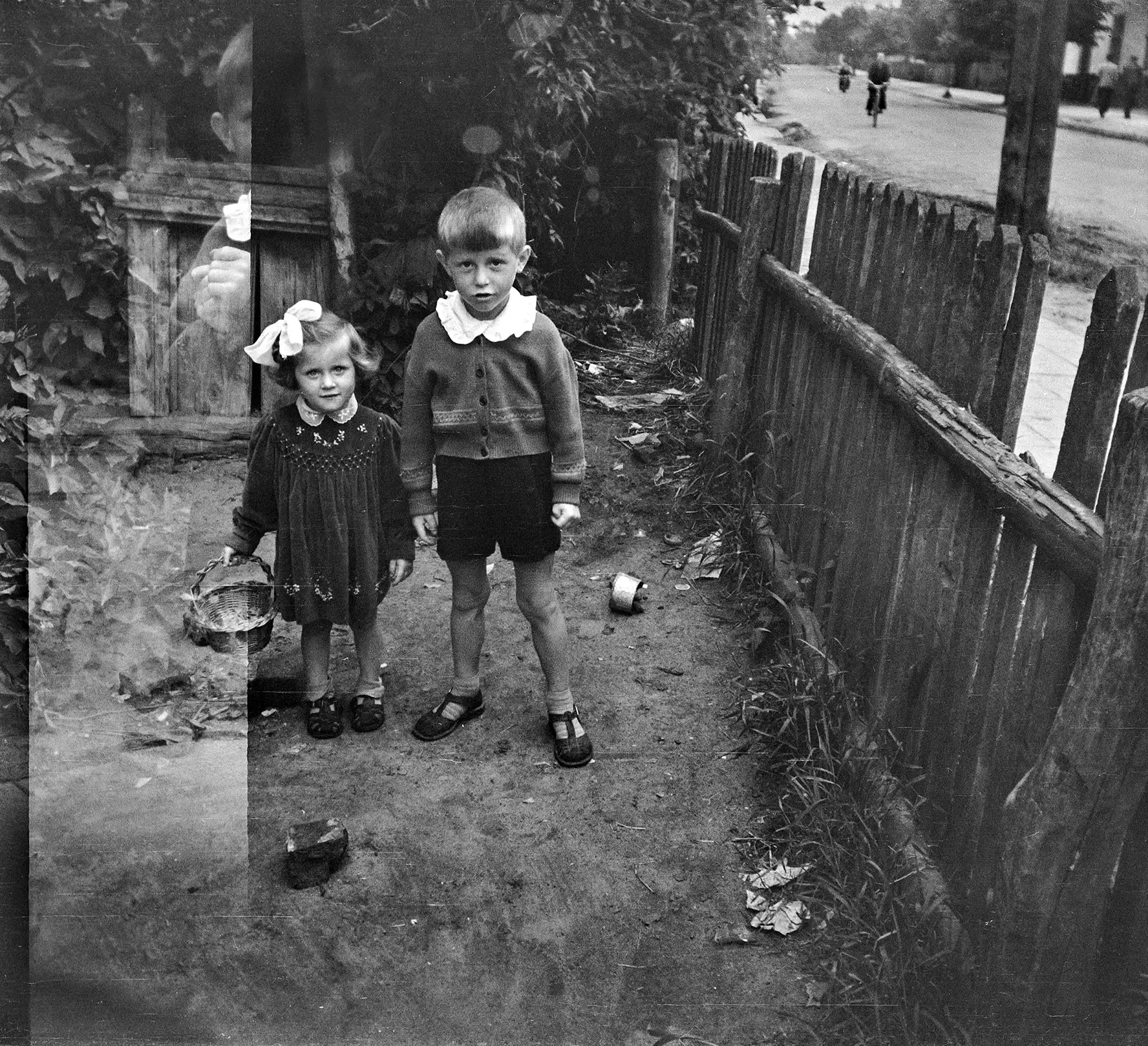 mała dziewczynka z koszyczkiem i mały chłopczyk pozujący na podwórku przy drewnianym płocie. Za płotem widoczna ulica