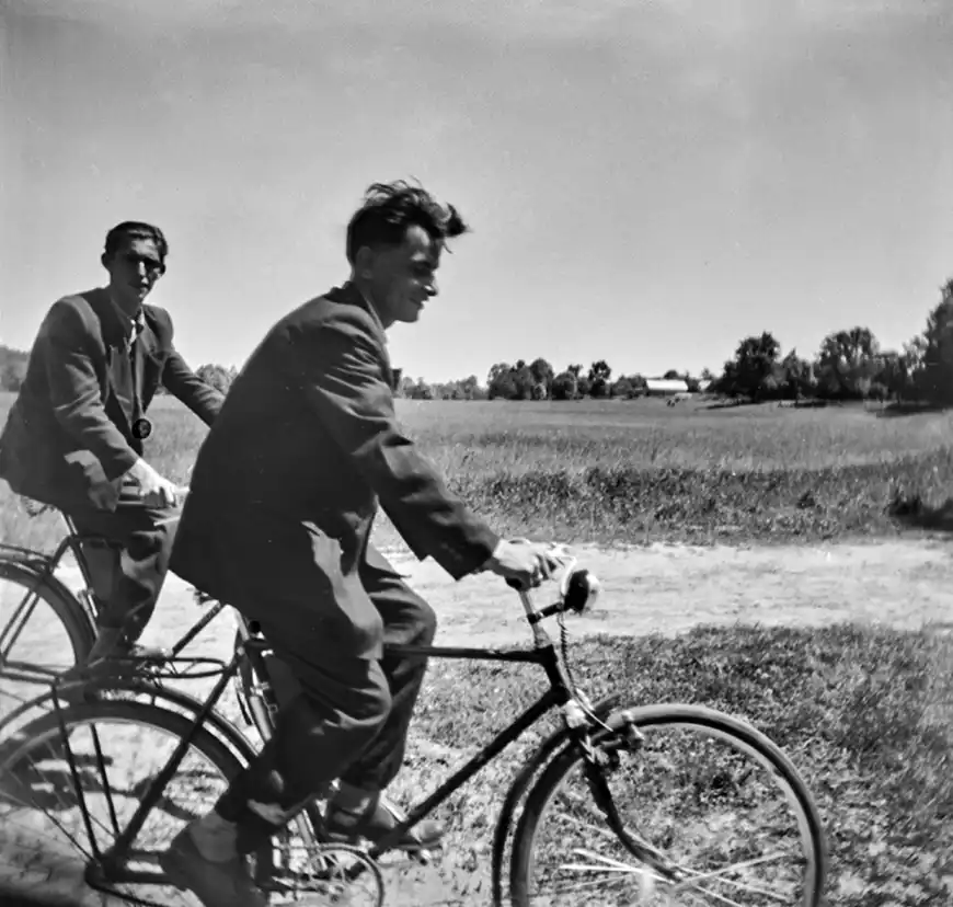 dwaj mężczyźni w marynarkach jadący na rowerach po ścieżce przez łąkę