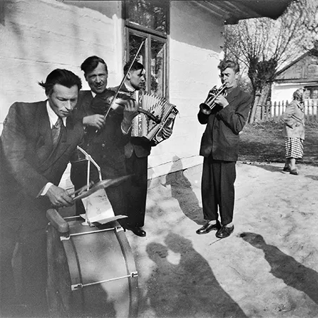 mężczyźni grający na perkusji, skrzypcach, akordeonie i trąbce przed drewnianym domem
