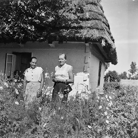 dwie kobiety wśród traw i kwiatów stojące na tle wiejskiej chaty krytej strzechą