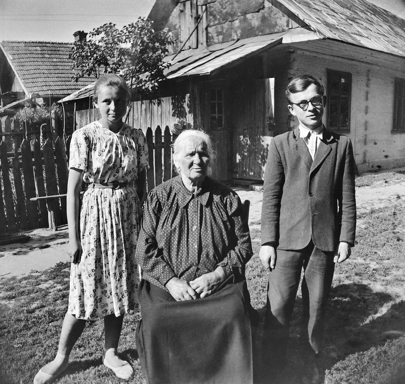 kobieta siedząca na podwórku przed wiejską chatą, po obydwu jej stronach młoda kobieta i młody mężczyzna