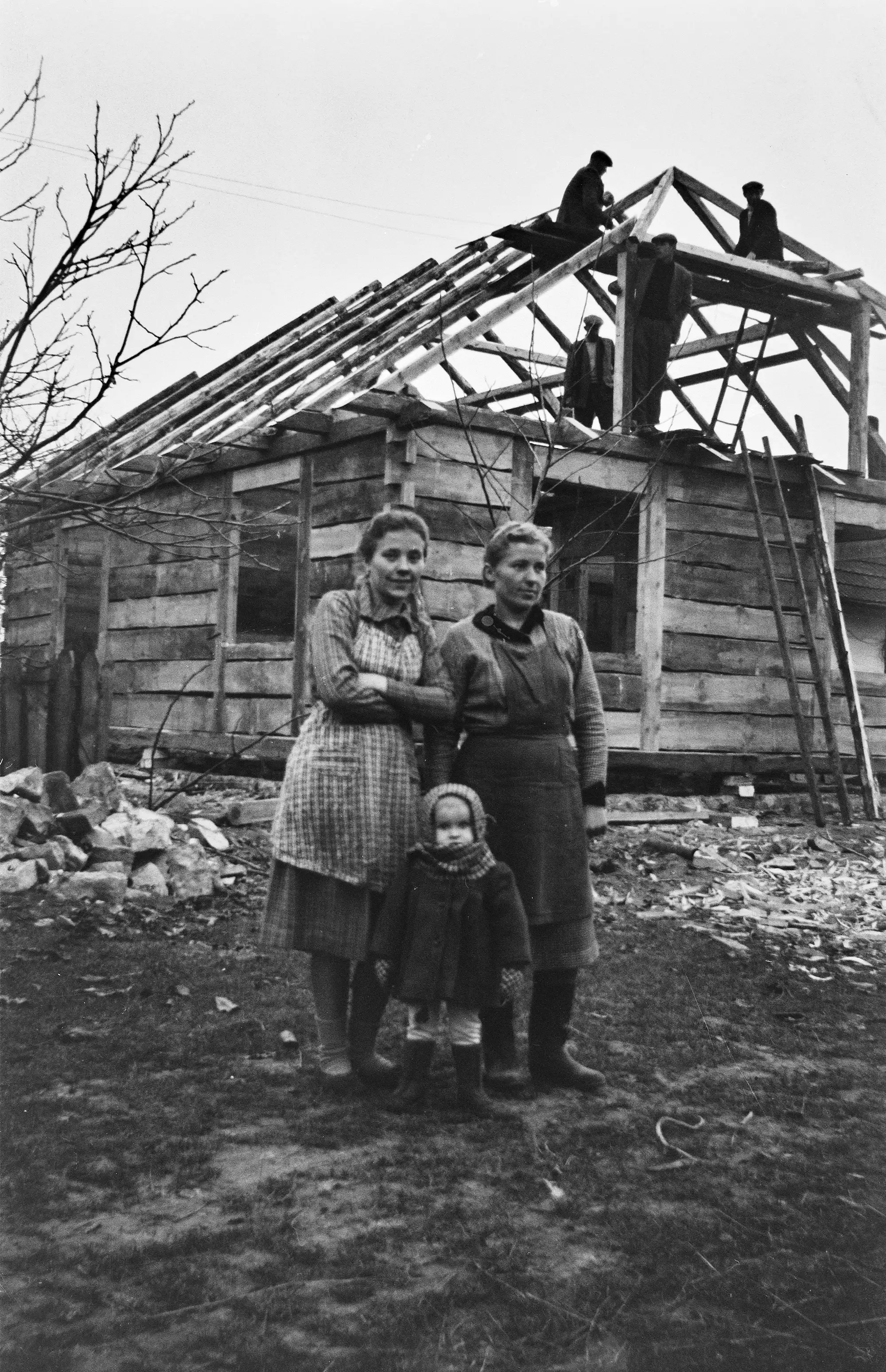 dwie kobiety i dziecko stojące na podwórzu, w tle budowa konstrukcji drewnianego domu, na konstrukcji stoją trzej mężczyźni