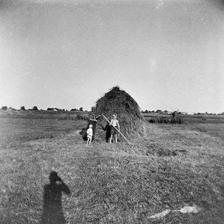 dalekie pole a na nim snop siana i dwójka dzieci i kobieta z grabiami, widać cień fotografa